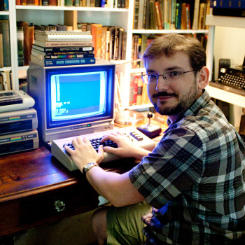 A bearded, bespectacled man sitting at an old computer but looking at the camera