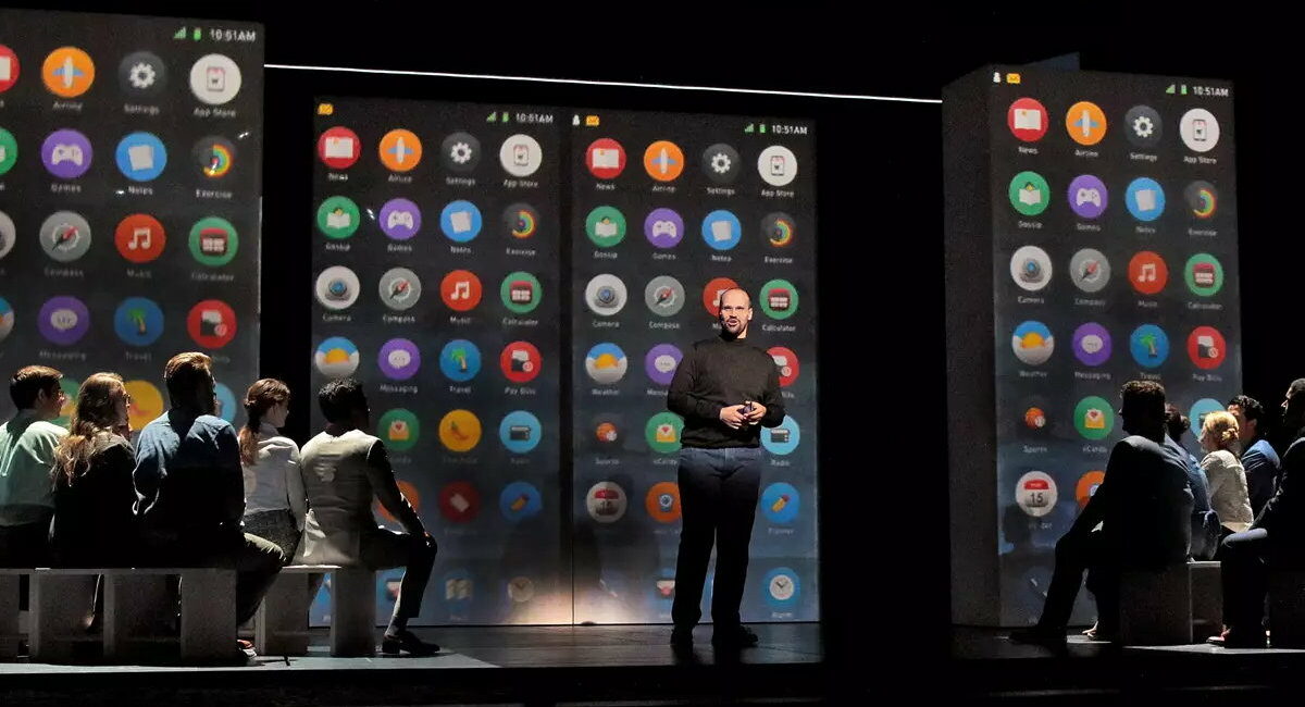 A theatrical stage with projects of iPhone displays in the background as one man stands in front of a seated audience