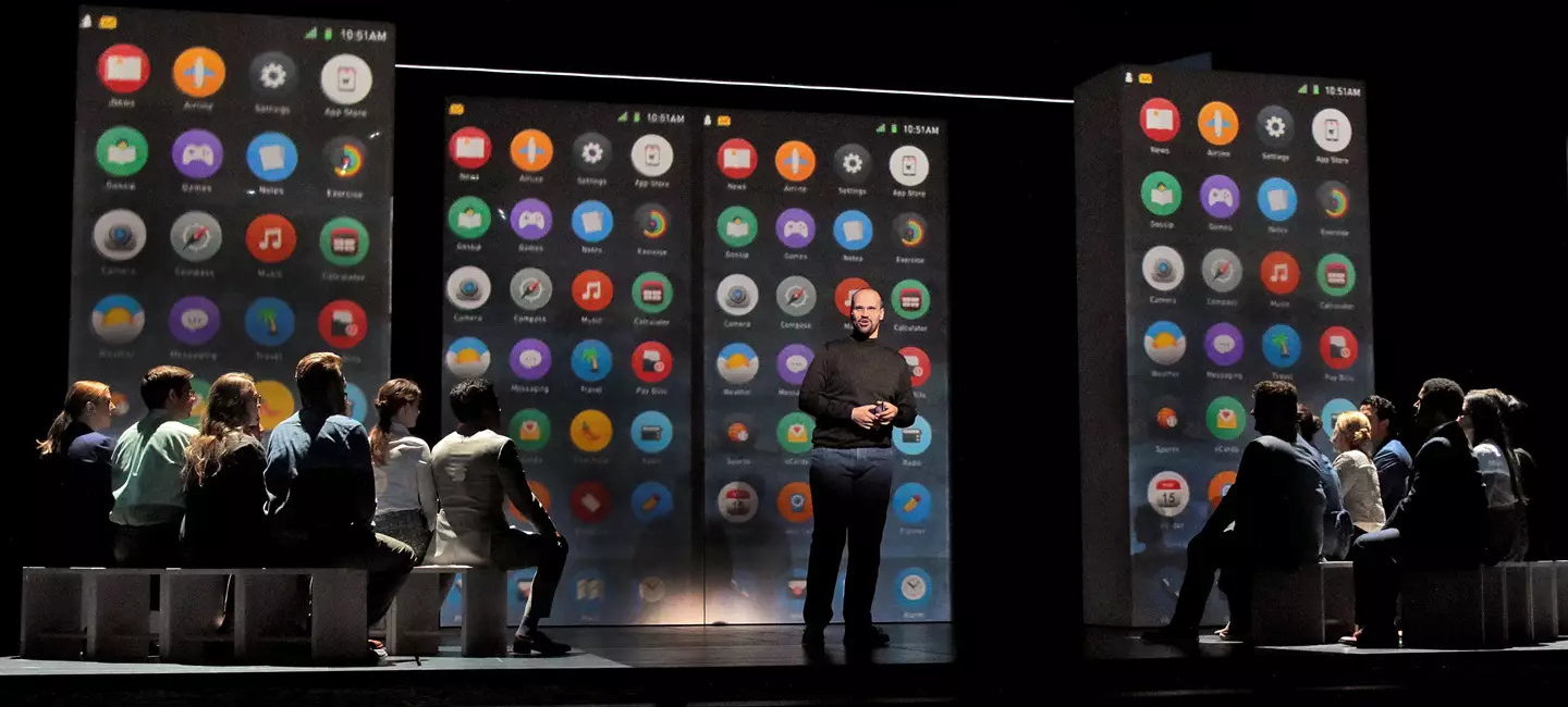 A theatrical stage with projects of iPhone displays in the background as one man stands in front of a seated audience
