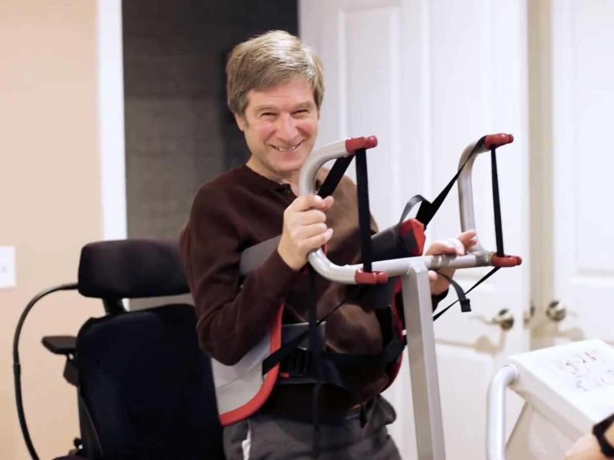 A smiling Bruce Rosenblum stands up with assistance during a physical therapy session.