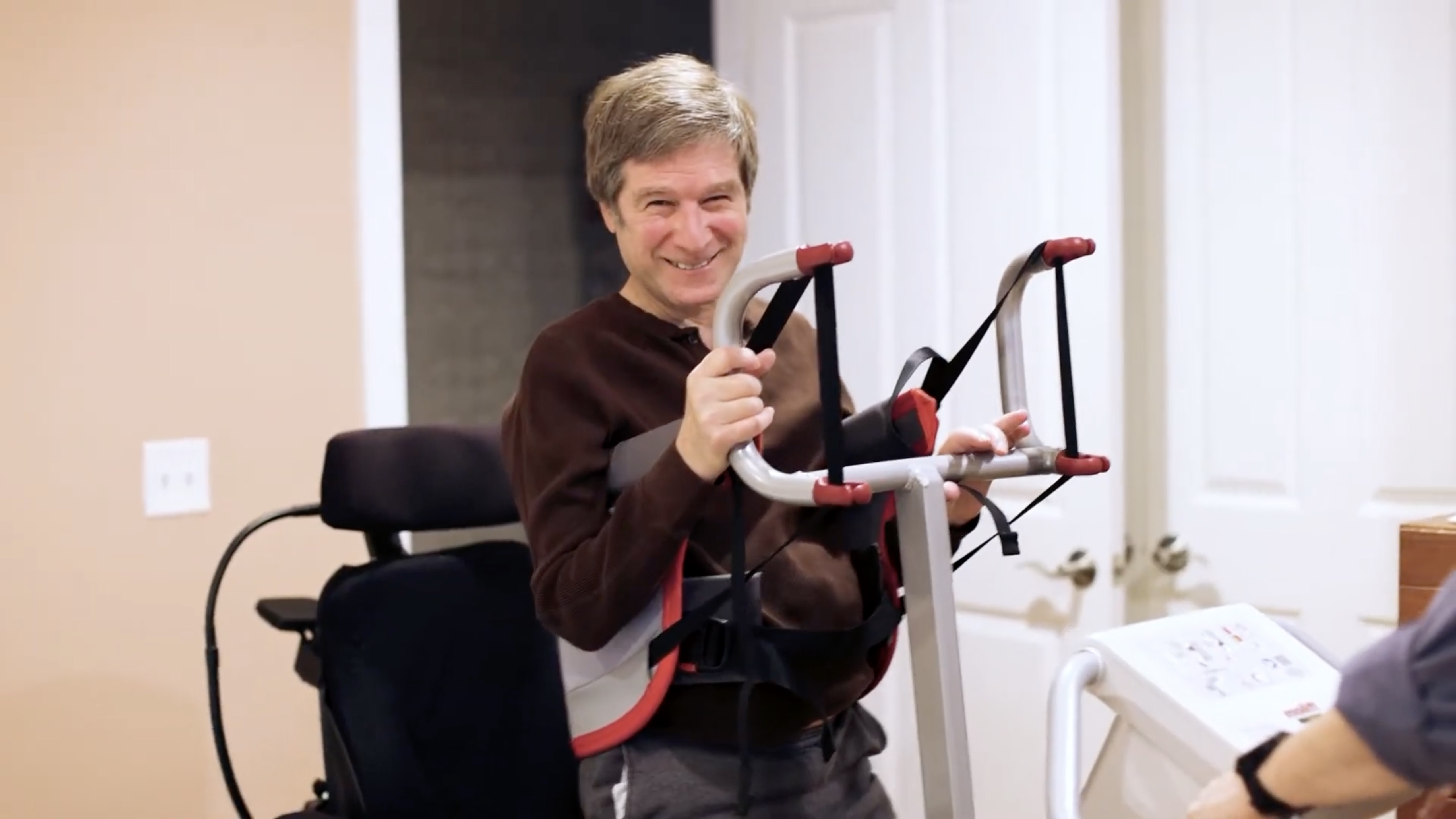 A smiling Bruce Rosenblum stands up with assistance during a physical therapy session.