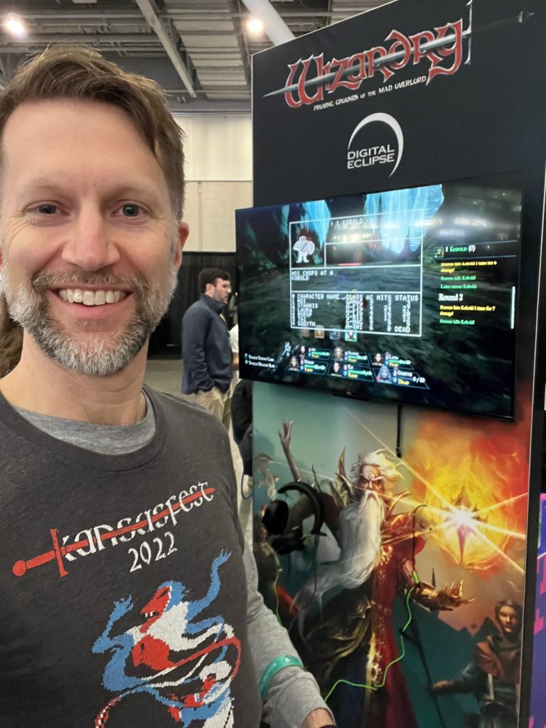 Ken wearing a Wizardry shirt from KansasFest 2022 in front of a Wizardry demo station at PAX East